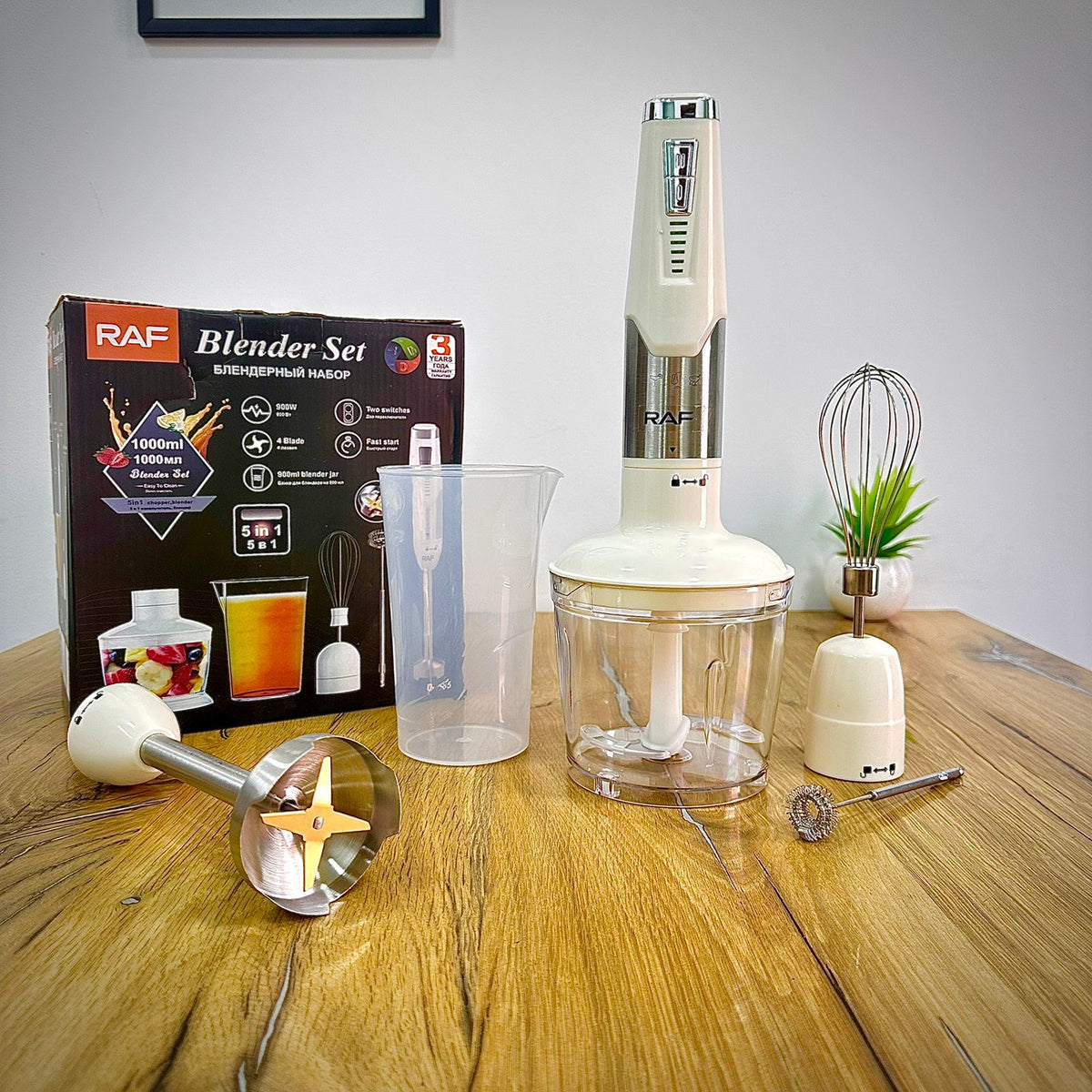 RAF Blender Set displayed on a wooden table with its retail box, clear plastic pitcher, immersion blender, whisk attachment, and small metal accessory. The set is photographed indoors against a plain light wall.