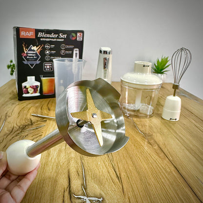 A stainless steel high-speed blender blade attachment with a white plastic handle is held by a hand in the foreground, with a boxed blender set and other blender components arranged on a wooden tabletop against a light gray wall.